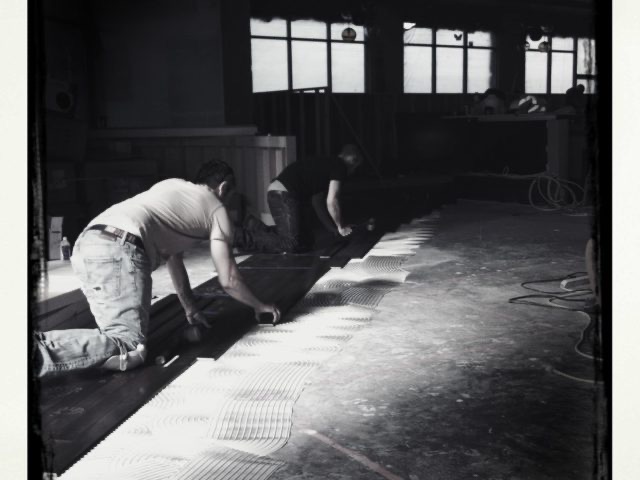 Installers laying dark plank flooring over trowel-spread adhesive in a commercial space